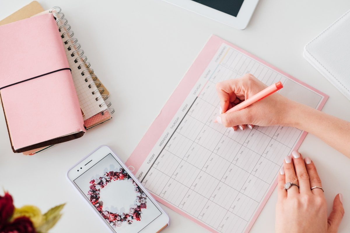 A woman's hand writing on a pink notebook next to a cell phone, determining the best time to post on social media.