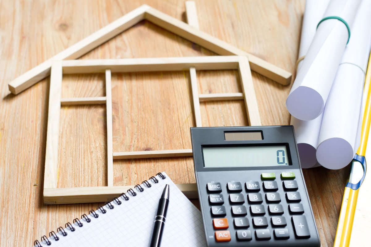 A calculator, pen, and house plans are neatly arranged on a wooden table in front of a home improvement business signage.