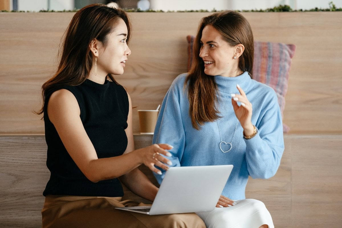 Two asian women discussing the content for their office's "About Us" page while using a laptop.