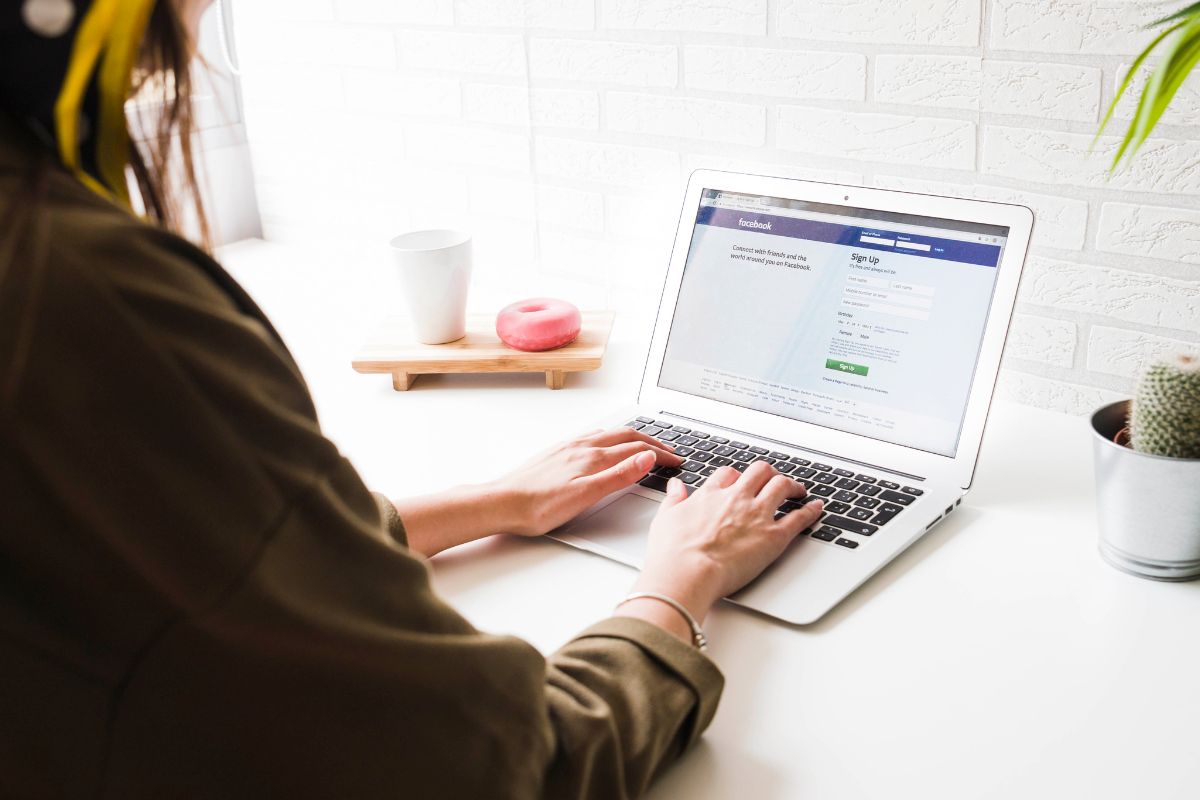 How to Create a Facebook Business Account: A Step-by-Step Guide 1 A woman wearing headphones using a laptop with a "how to create a Facebook business account" guide, sitting at a white desk with a coffee cup, apple, and potted plant.