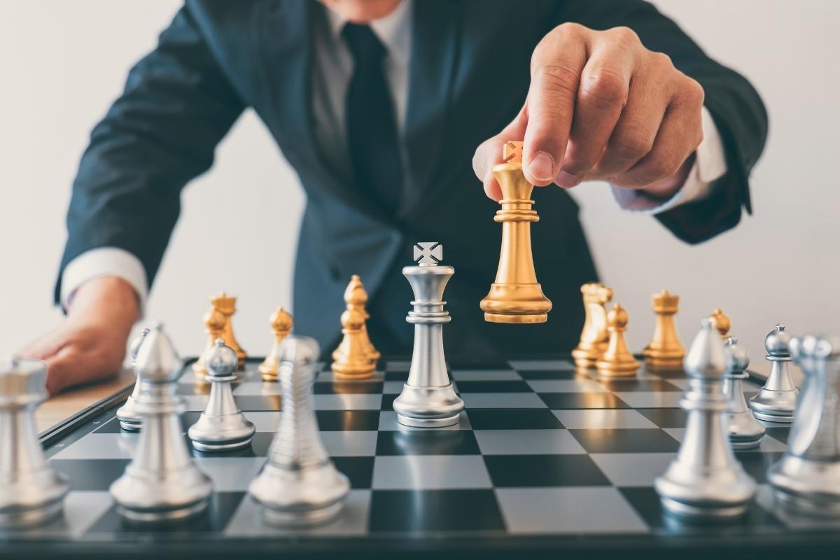 What Are Marketing Tactics & Their Benefits? 7 A businessman strategically places a gold piece on a chess board, utilizing marketing tactics.