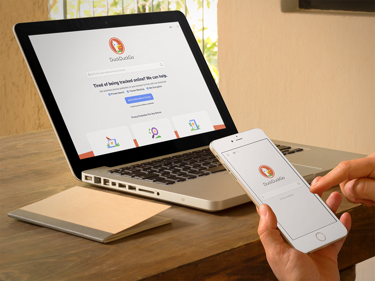 A person multitasking with a phone and laptop on a table while searching on DuckDuckGo.