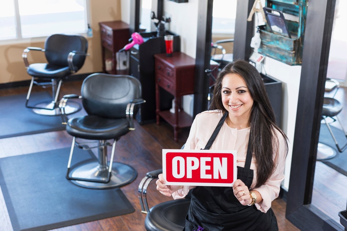 A woman implementing a beauty salon marketing plan while holding an open sign in her hair salon.