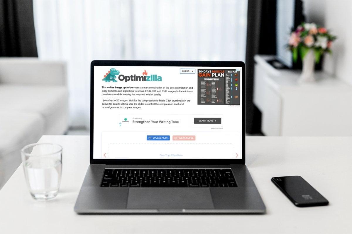 A laptop is sitting on a table in front of a window while utilizing an online image compressor like Optimizilla.