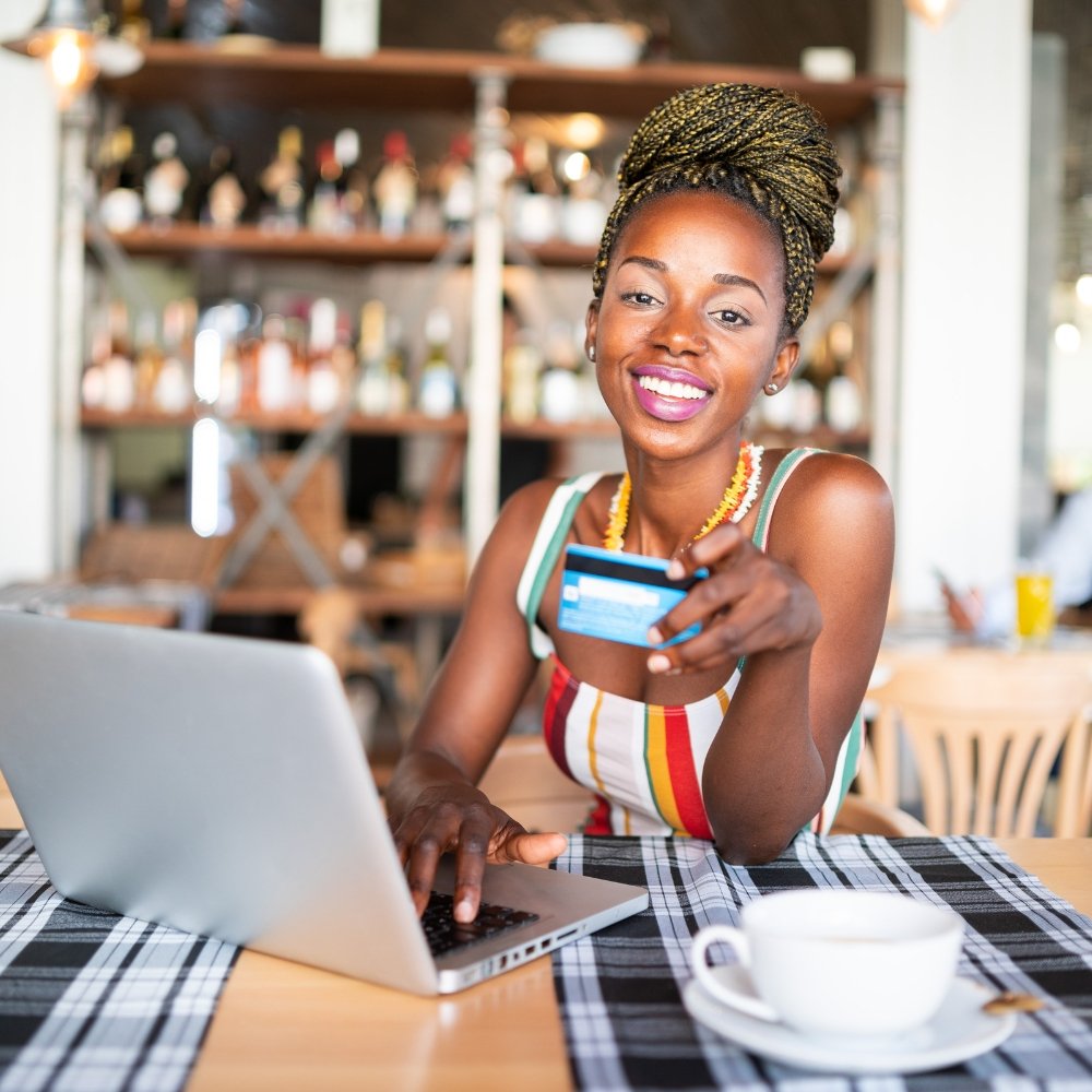 A young woman testing a WooCommerce website design on her laptop, while holding a credit card in a cafe.