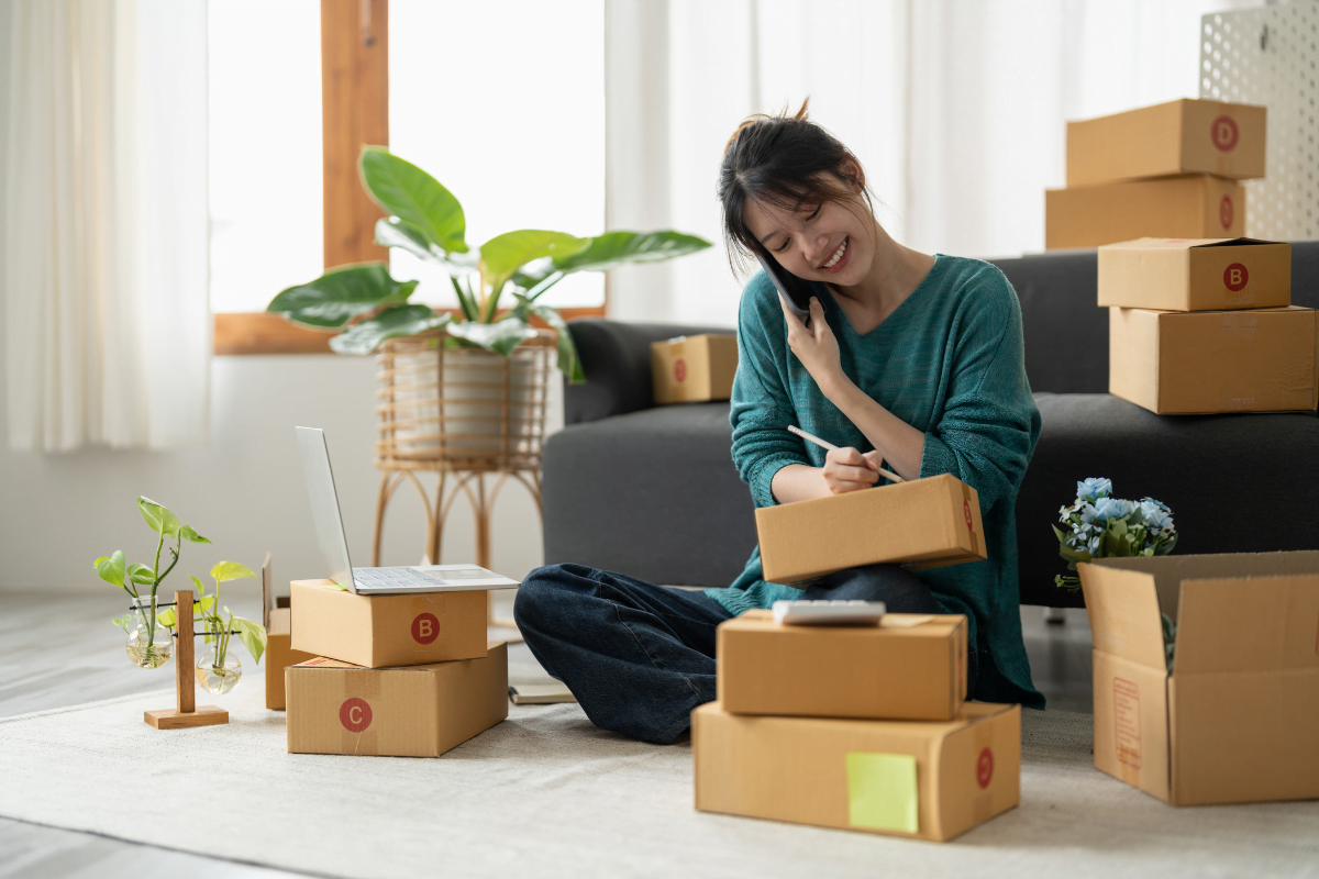 10 Game-Changing Strategies To Skyrocket Your eCommerce Business Success 7 A woman sitting on the floor with boxes, managing her ecommerce business from home.