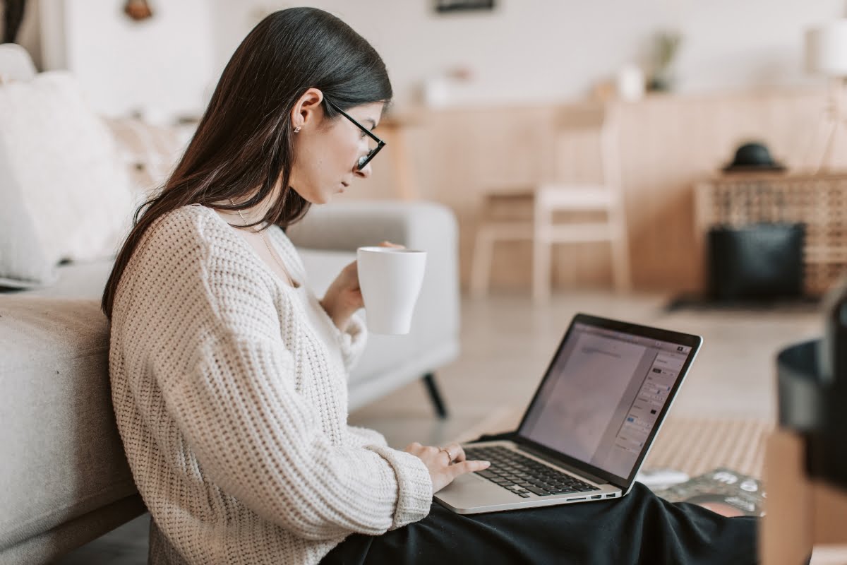 A woman lounging next to the couch with a laptop and a cup of coffee, fixing low quality content on her website.