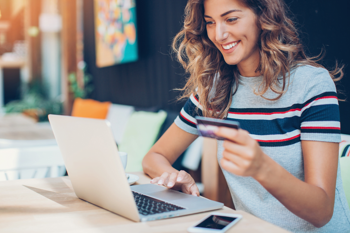 Boost E-Commerce Sales With WooCommerce Gift Cards Plugin 4 A woman is using WooCommerce gift cards to make a purchase while sitting at a table with a laptop and a credit card.