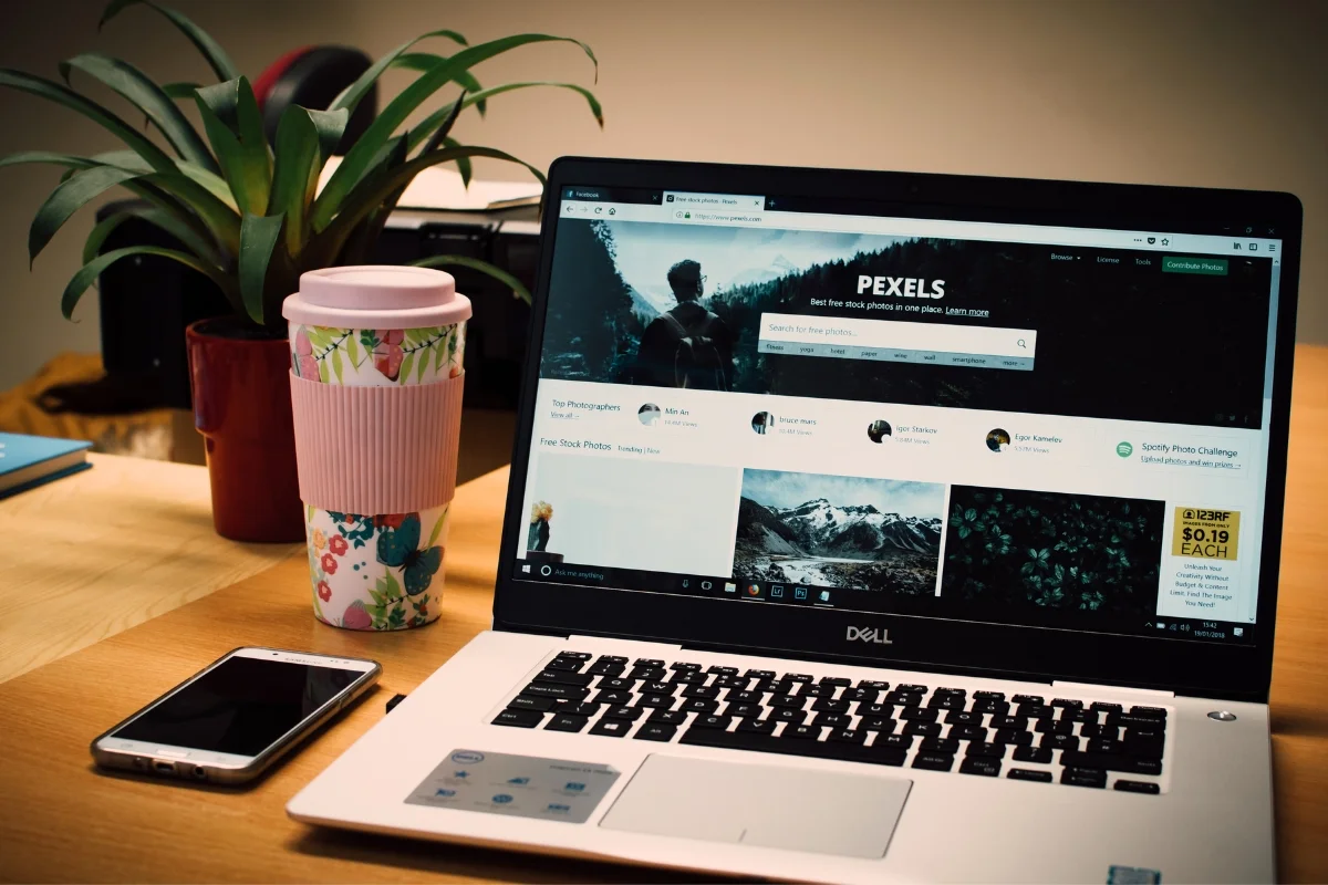 A laptop is sitting on a desk next to a cup of coffee and a plant in website photos.