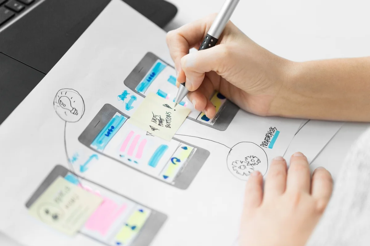 How to Create Wireframes: A Beginner's Guide for Stunning Designs 7 A person sketching how to create wireframes for a mobile app interface on paper with annotations and icons.