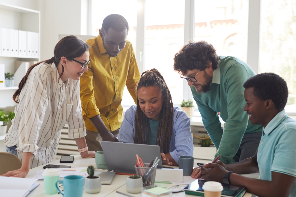 A diverse team of five outsourced marketing professionals collaborating around a laptop in a well-lit office environment.