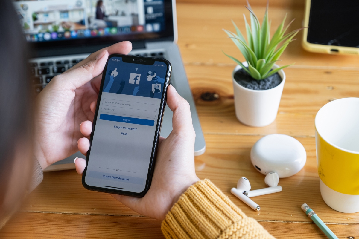 How to Improve Your Facebook Business Page With These Expert Tips 2 Person holding a smartphone with a Facebook business page open, surrounded by a laptop, earbuds, a notepad, and a cup on a wooden desk.