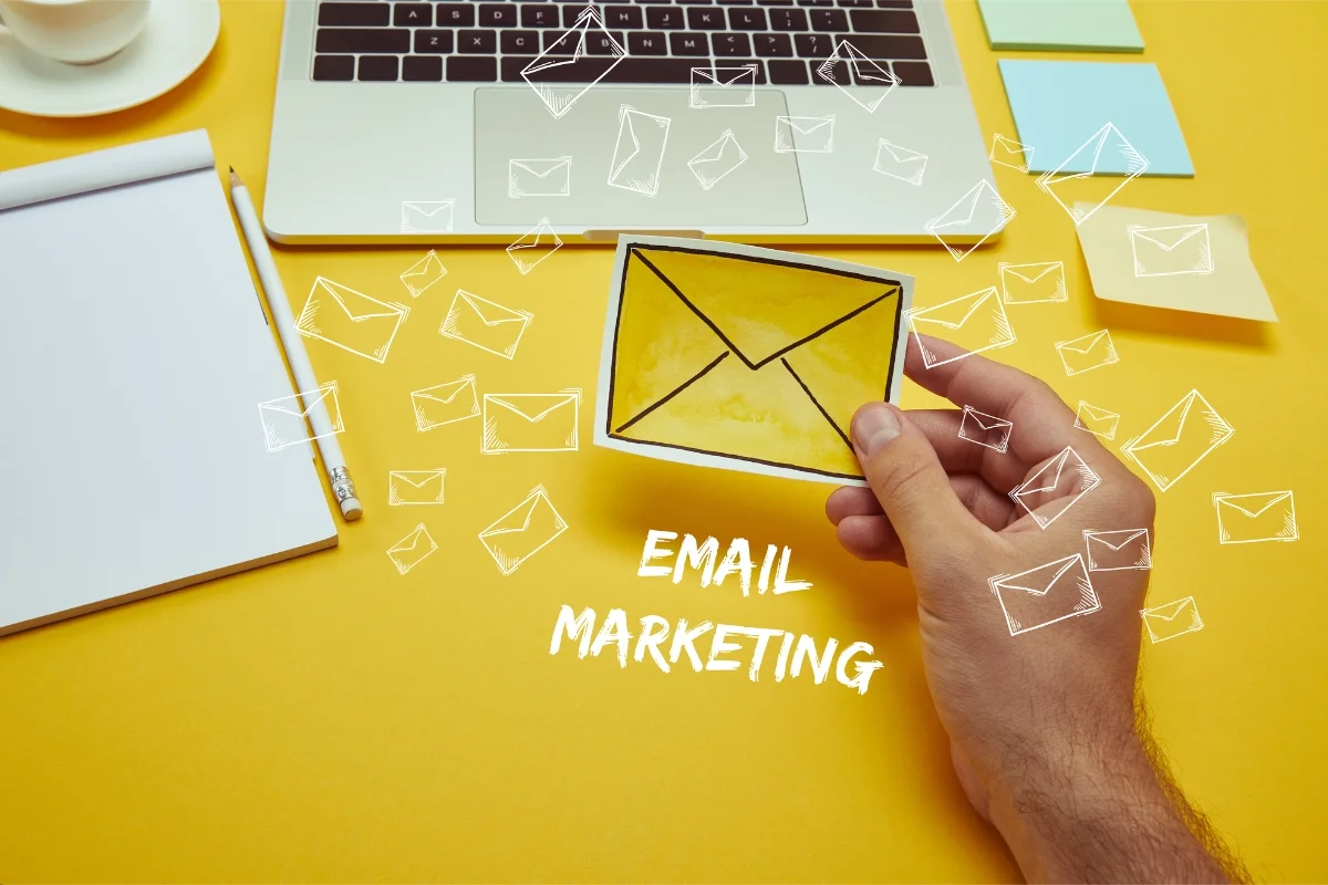 Email Targeting: Master the Art of Personalized Messaging 4 A hand holds a yellow envelope in front of a laptop and a notepad, with the words "Email Marketing" written below. Various envelope icons float in the background, emphasizing effective email targeting strategies.