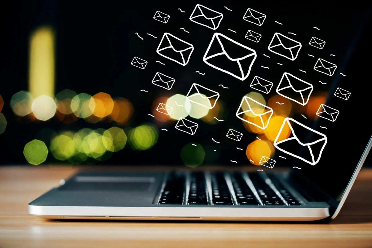 Email Targeting: Master the Art of Personalized Messaging 8 A laptop on a desk with illustrated email icons emerging from the screen, indicating email targeting and communication. Faint city lights are blurred in the background.