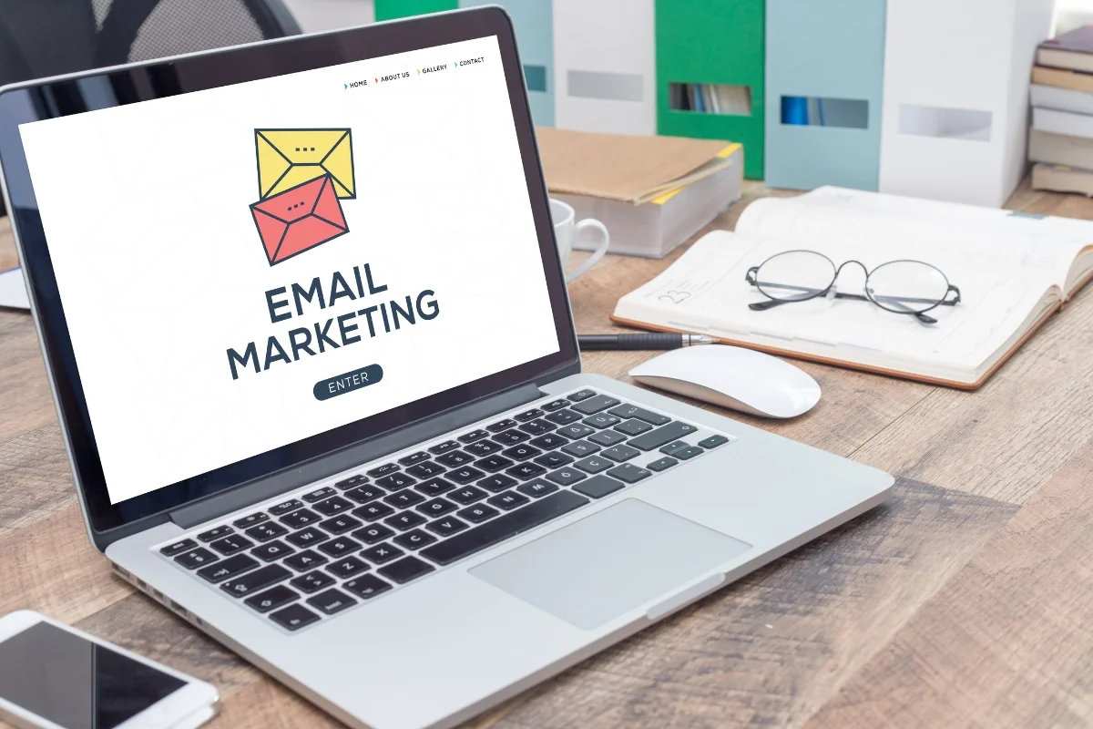Email Targeting: Master the Art of Personalized Messaging 2 A laptop displaying an "Email Marketing" screen sits on a wooden desk cluttered with a mouse, notebook, glasses, smartphone, and various office supplies, all essential tools for effective email targeting.