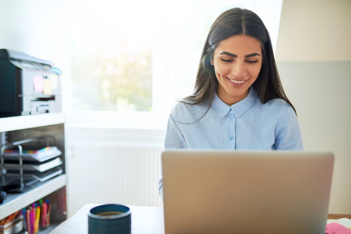 What Is AB Testing in Marketing: Unlocking Data-Driven Success 5 A woman sits at a desk with a smile, working on a laptop in a bright, sunlit room. A cup and printer are visible in the background as she possibly researches what is AB testing in marketing.
