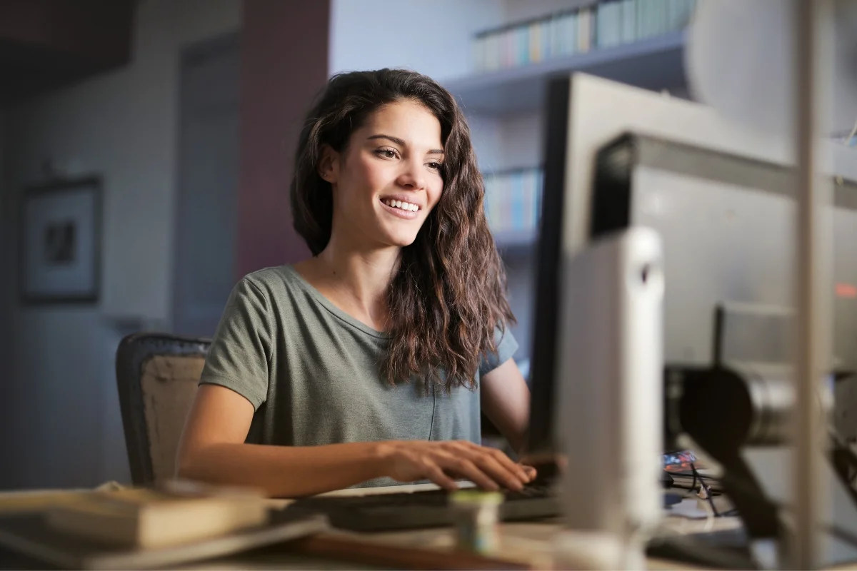 What Is AB Testing in Marketing: Unlocking Data-Driven Success 2 A woman with long hair and a green shirt is smiling while using a desktop computer, possibly researching what A/B testing in marketing is. Bookshelves are visible in the background.