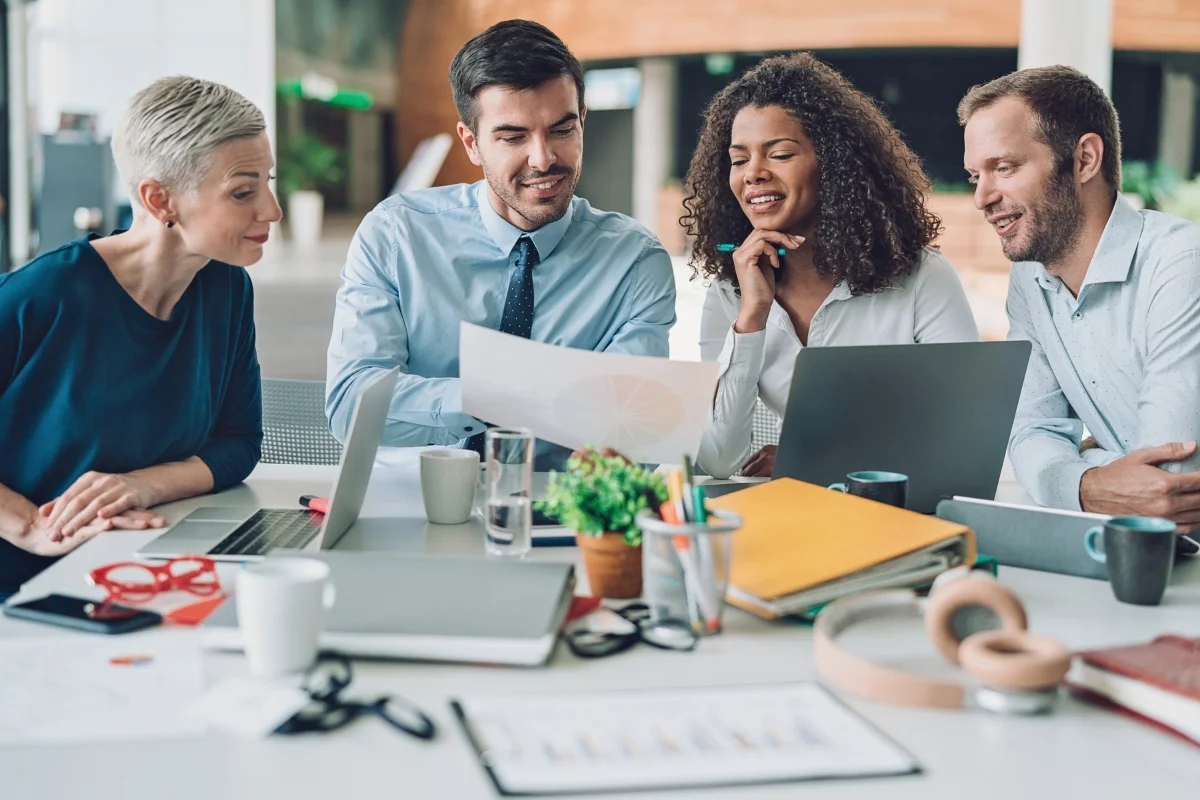 What Is Hyperlocal Marketing: Targeting for Local Business Growth 8 Four people sitting at a desk with laptops and papers, engaged in a discussion about what is hyperlocal marketing and reviewing a document in an office setting.