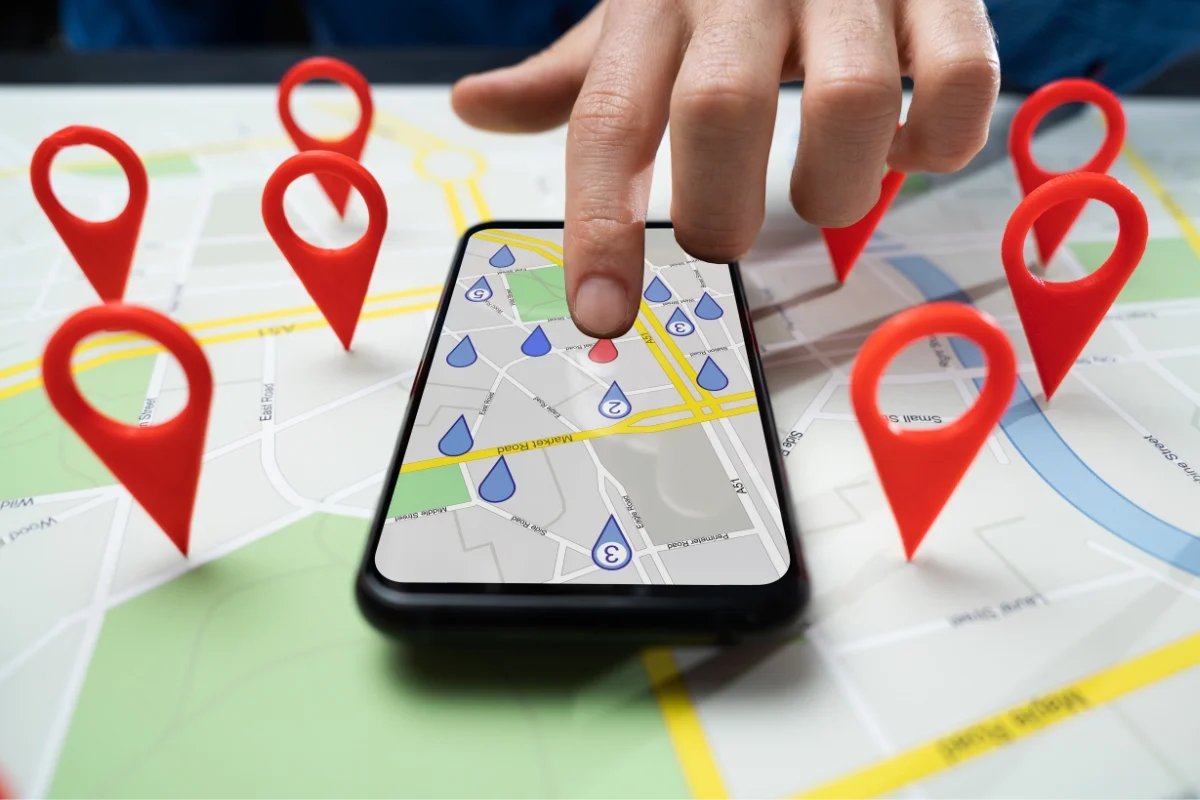What Is Hyperlocal Marketing: Targeting for Local Business Growth 2 A hand points at a smartphone displaying a map with multiple location markers set on a physical map. Red location markers are also placed on the actual map around the phone, illustrating what is hyperlocal marketing in action.