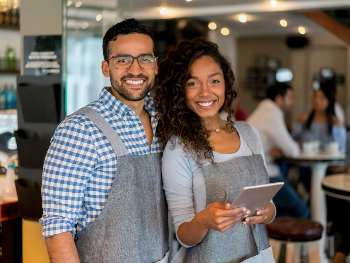 Two smiling individuals, one holding a tablet, are wearing aprons and standing in what appears to be a busy caf&eacute; or restaurant, possibly discussing citation building for their online menu.