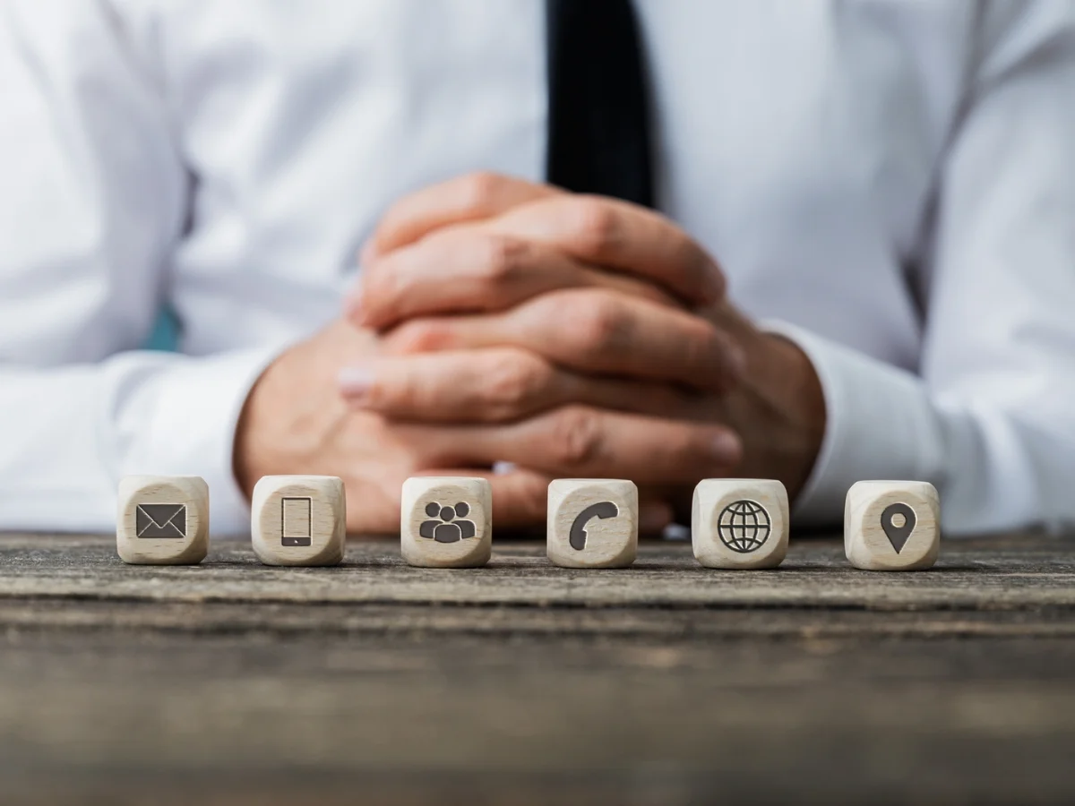 A person with hands folded sits behind a table with six wooden blocks displaying icons: envelope, smartphone, group of people, phone, globe, and location pin &mdash; tools essential for effective citation building.