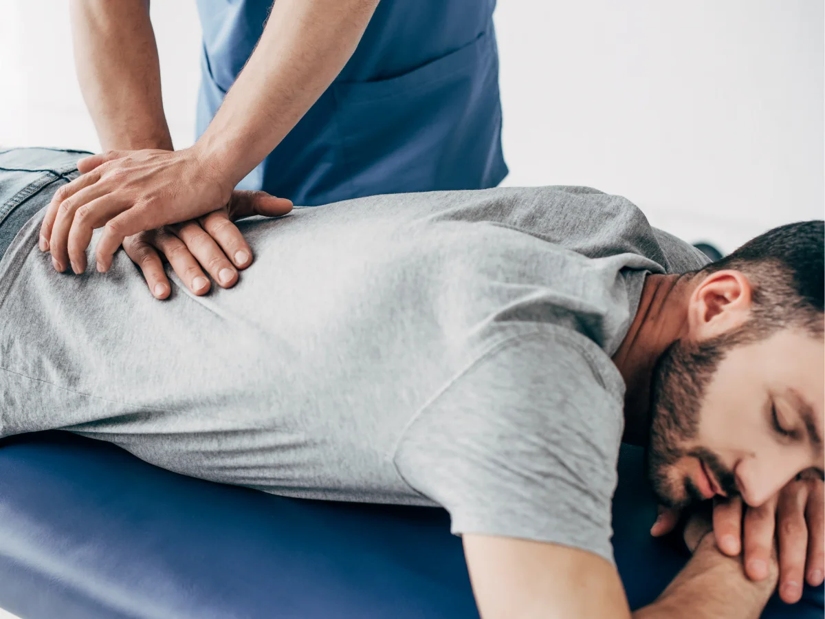 A person in a gray shirt lies face down on a blue table while another person performs a back massage.