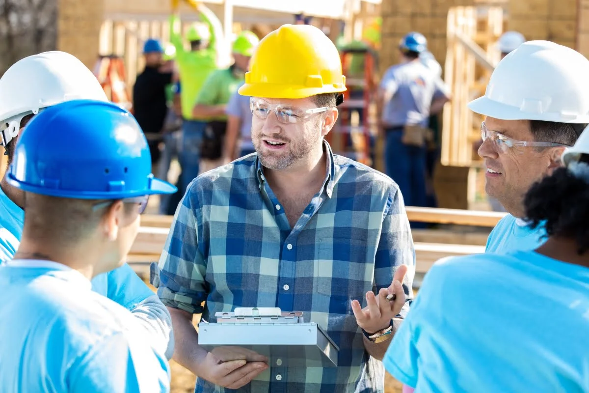Construction CRM Software 27 A construction manager in a yellow hard hat holds a clipboard and speaks to a group of workers wearing blue hard hats at a building site. Other workers and construction activities are visible in the background.