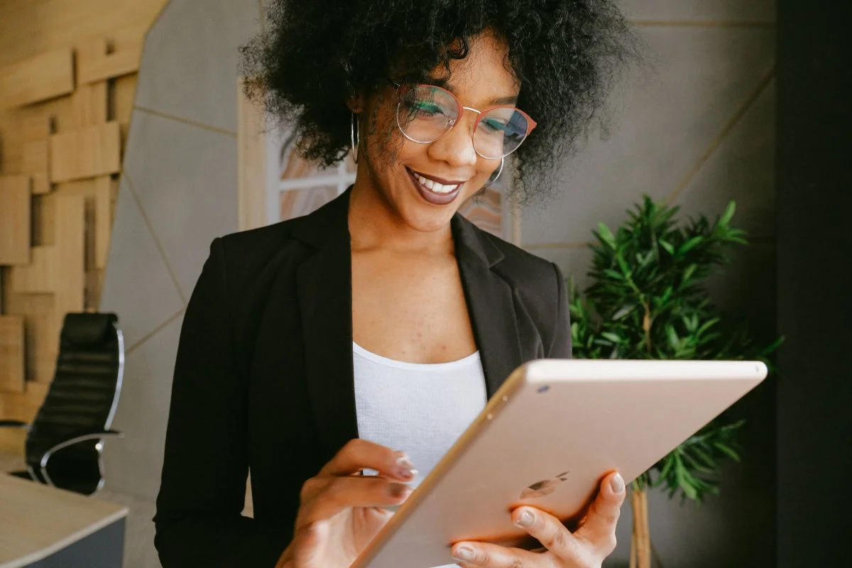 Maximize Efficiency: CRM Integration With Business Automation 5 A person in a blazer and glasses is smiling and using a tablet in an office setting with a chair, plant, and CRM integration tools in the background.