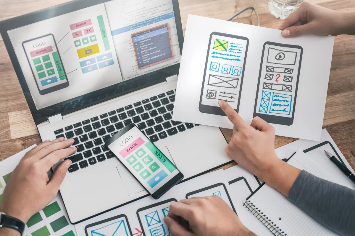 People working collaboratively on a mobile app design project, discussing how website design affects sales, with printed wireframes, a smartphone with a design prototype, and a laptop displaying a coding interface.
