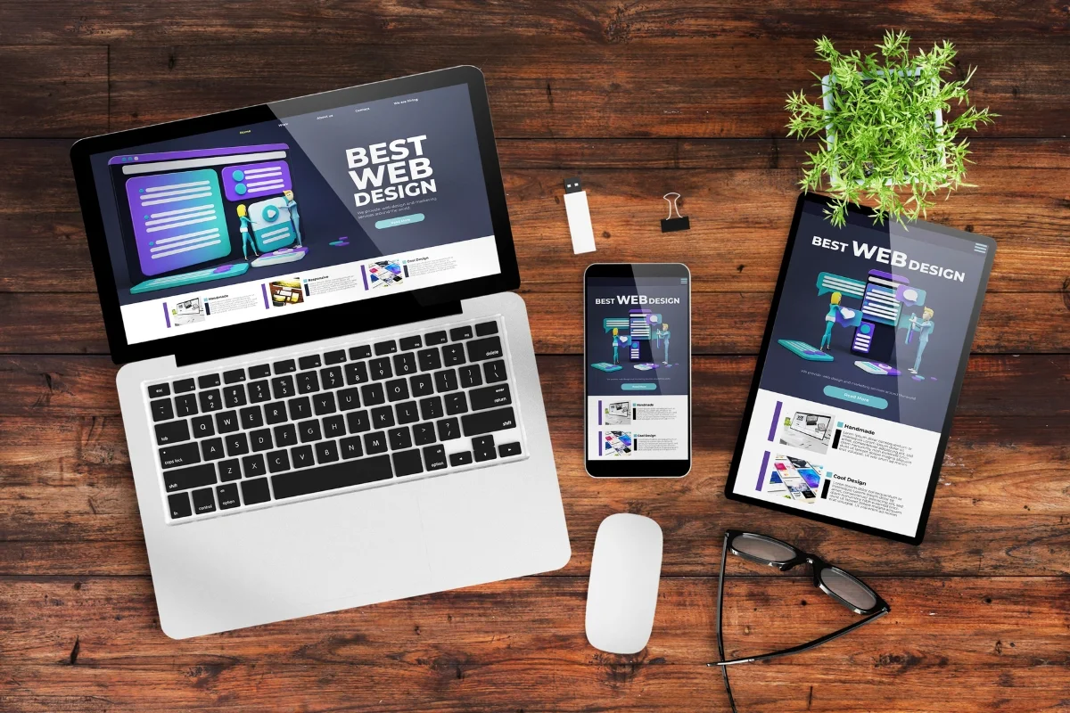Laptop, smartphone, and tablet displaying a web design website on a wooden desk with a mouse, reading glasses, USB drive, and a small plant&mdash;every element meticulously placed to answer the question: how does website design affect sales?
