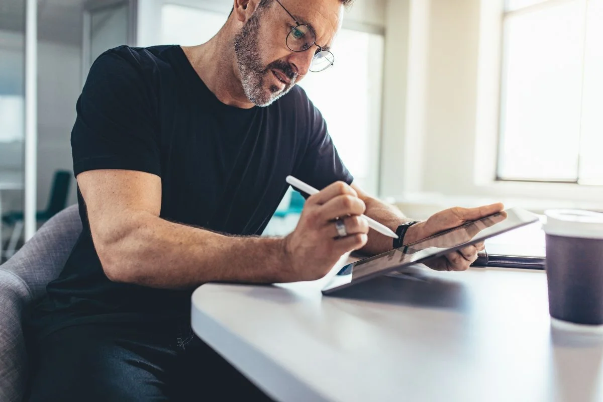 Man in glasses and a black t-shirt sitting at a table, writing on a tablet with a stylus. A coffee cup is placed nearby as he researches email marketing automation tools.