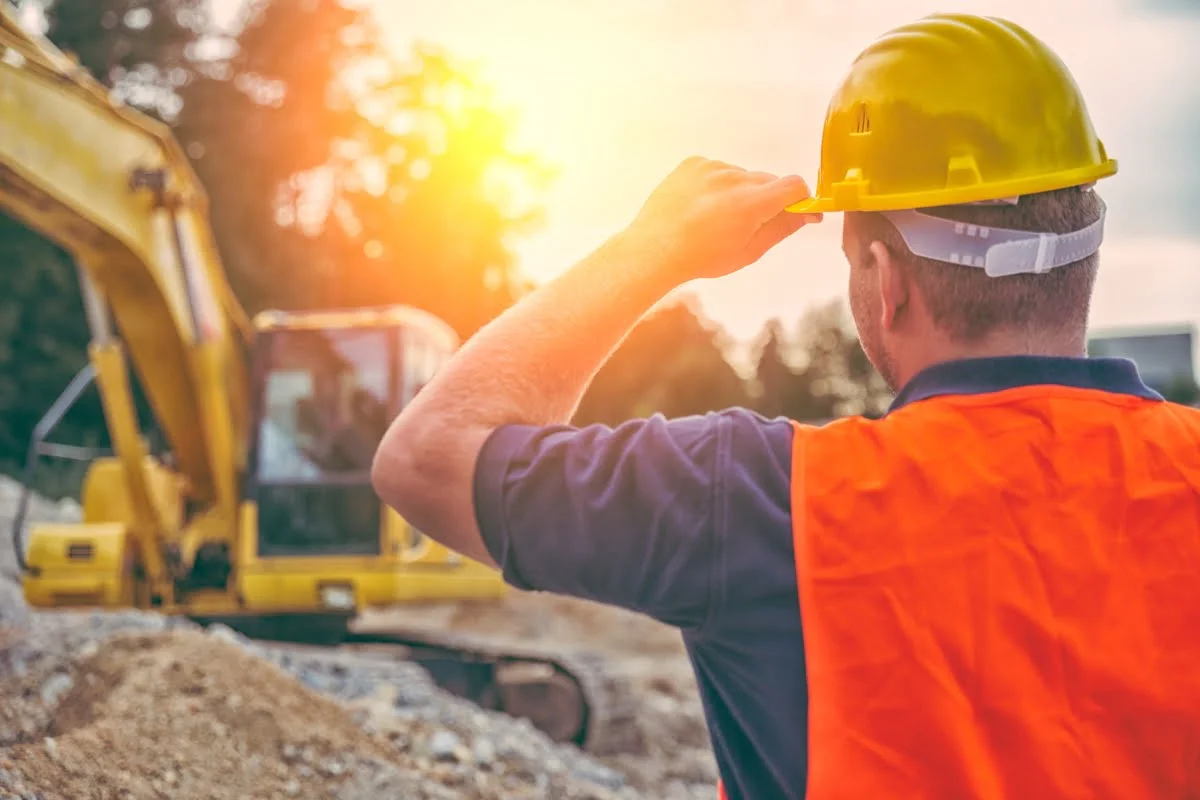 Construction CRM Software 28 A construction worker wearing a yellow hard hat and orange vest observes an excavator at a construction site during sunset.