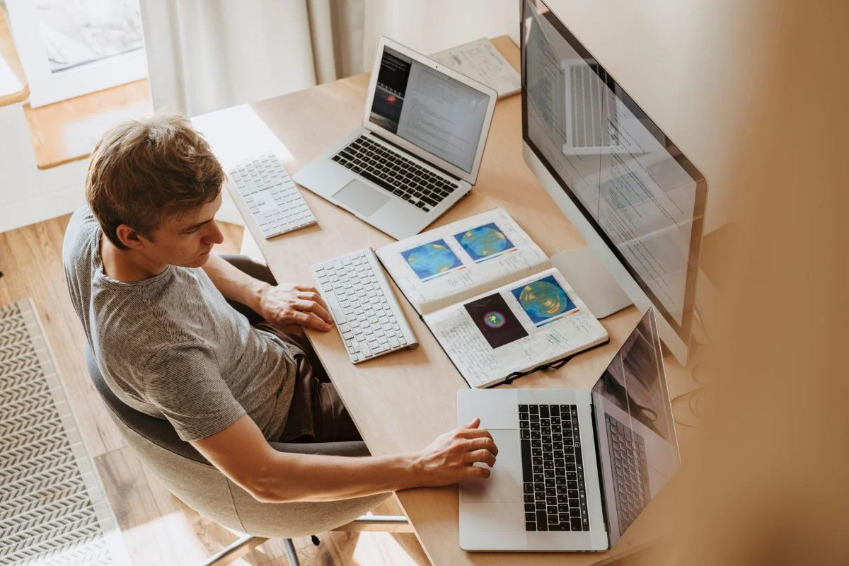 Maximize Efficiency: CRM Integration With Business Automation 3 A person sits at a desk using multiple laptops and a large monitor, seamlessly managing tasks with crm integration tools, with open books and a notebook in front of them.