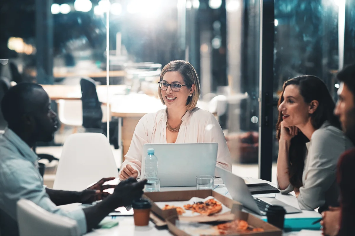 A group of four people sits around a table in an office, engaged in discussion about sales process automation. One woman is using a laptop, and there are open pizza boxes and drinks on the table.