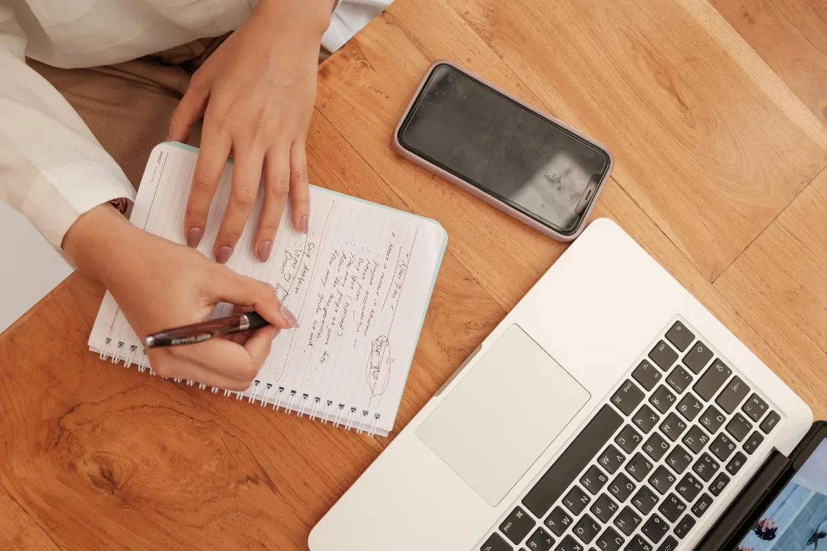 Enhance Your WordPress Site by Mastering Engaging Content Creation 3 A person engages in content creation, writing notes in a notebook on a wooden desk, with a smartphone and open laptop beside them.