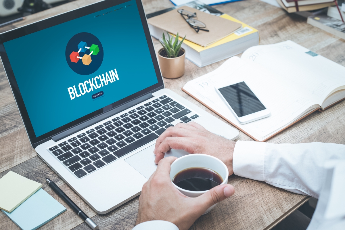 Blockchain in Digital Marketing: Potential Use Cases 2 Person using a laptop displaying the word "Blockchain" on screen, holding a cup of coffee at a desk with a notebook, pen, smartphone, and small plant—highlighting the role of Blockchain in Digital Marketing.