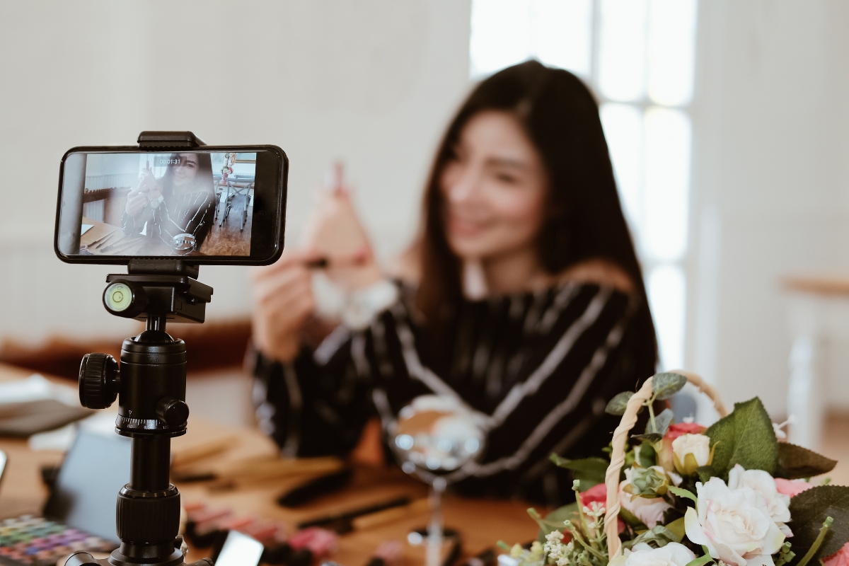 Blockchain in Digital Marketing: Potential Use Cases 4 A smartphone on a tripod records a woman at a table with makeup products and flowers, as she discusses an item—possibly sharing insights on Blockchain in Digital Marketing with her audience.