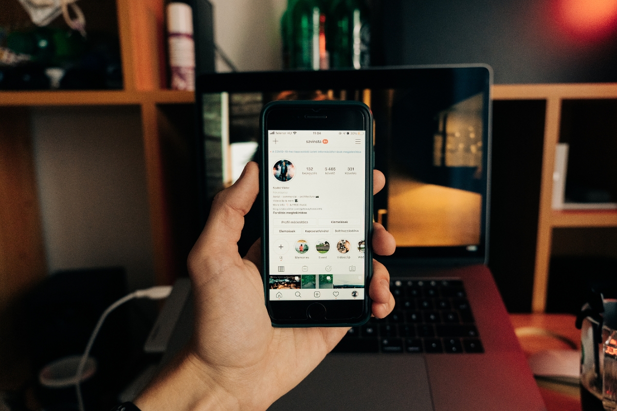 Blockchain in Digital Marketing: Potential Use Cases 5 A person holds a smartphone displaying an Instagram profile in front of an open laptop on a cluttered desk, showcasing the growing influence of blockchain in digital marketing strategies.
