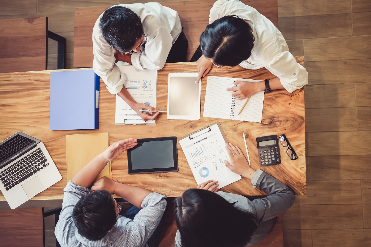 Blockchain in Digital Marketing: Potential Use Cases 6 Four people sit around a wooden table with laptops, tablets, documents, and a calculator, actively discussing business charts and notes—including strategies for using blockchain in digital marketing.