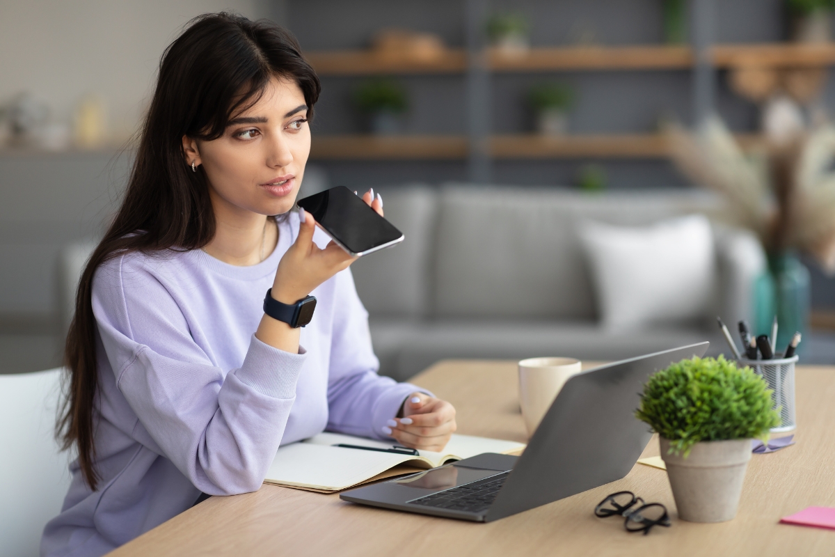 How Does Voice Search Work? A Beginner’s Guide to Smart Technology 2 A woman sits at a desk in her modern home office, using her smartphone’s voice function to ask, “How does voice search work?” while a laptop, notebook, and coffee cup sit in front of her.