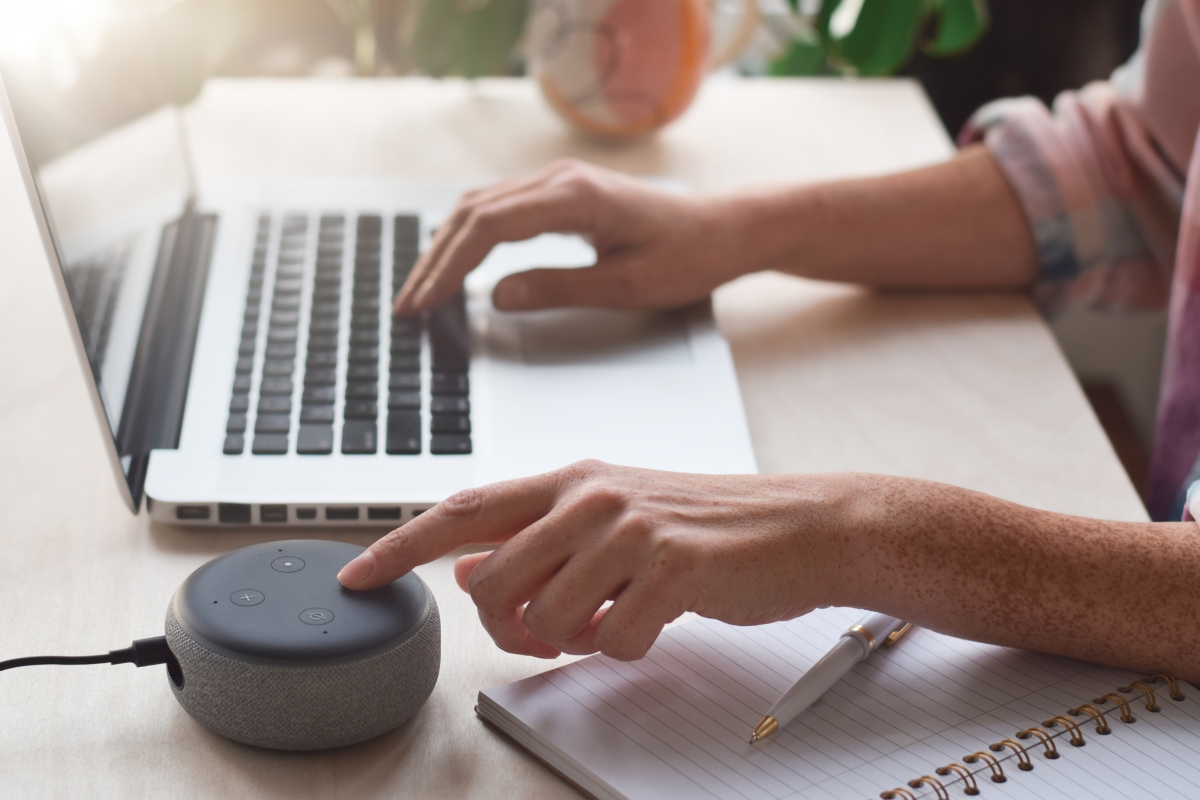 How Does Voice Search Work? A Beginner’s Guide to Smart Technology 3 A person works on a laptop, writes in a notebook, and interacts with a smart speaker on a desk, exploring how voice search works in daily tasks.