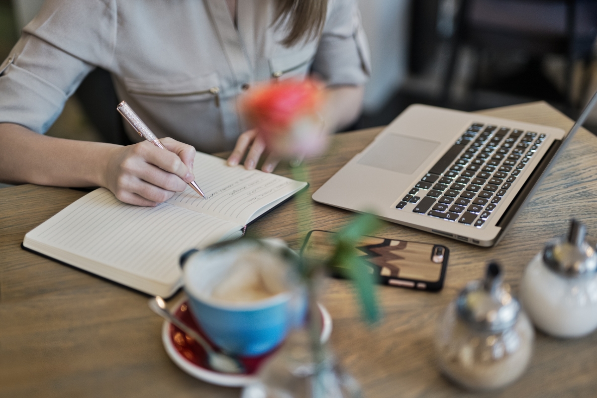How Does Voice Search Work? A Beginner’s Guide to Smart Technology 6 Person writing in a notebook next to an open laptop, a smartphone displaying "How does voice search work," and a cup of coffee on a wooden table.