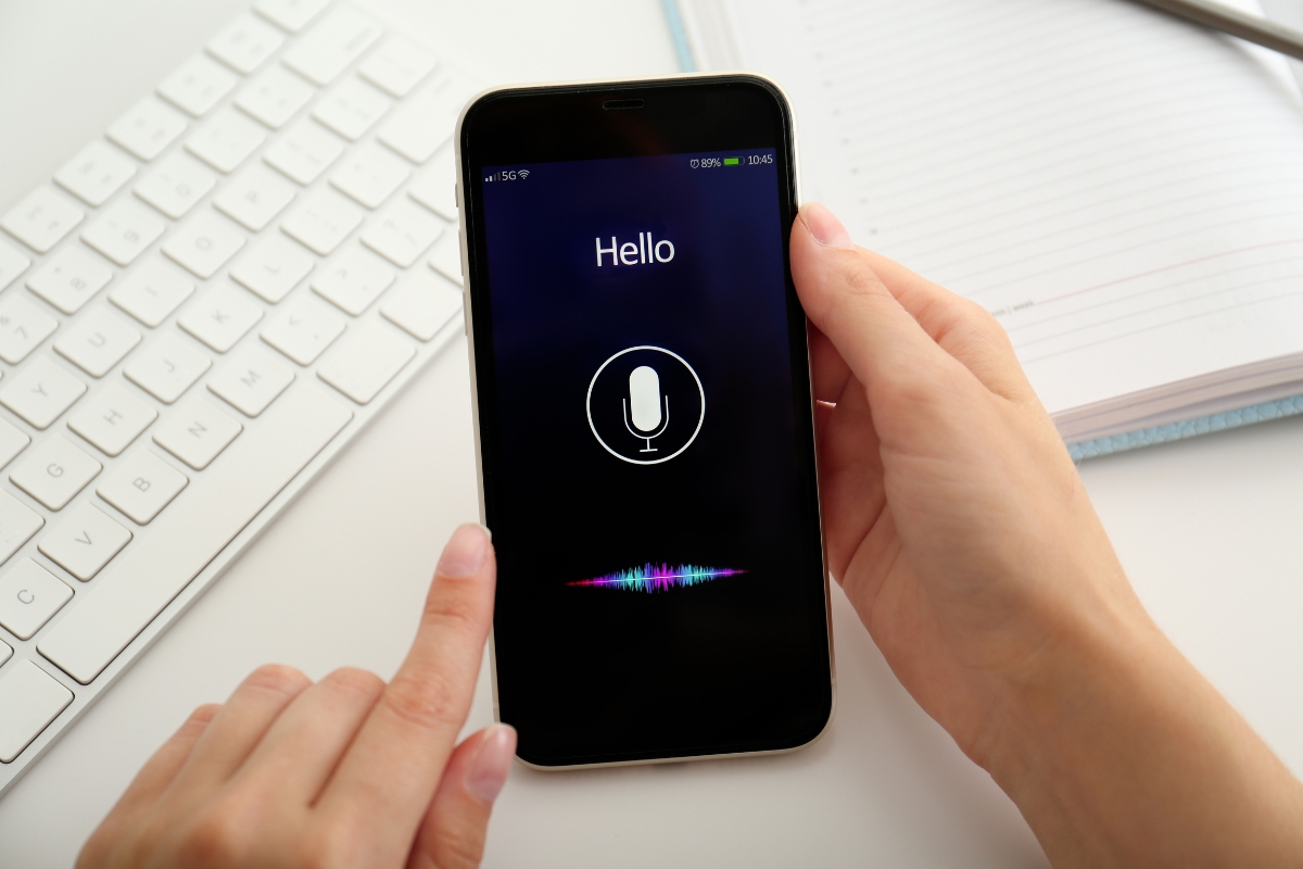 How Does Voice Search Work? A Beginner’s Guide to Smart Technology 7 A person holds a smartphone displaying a voice assistant interface with the word "Hello" and a microphone icon, illustrating how voice search works, next to a keyboard and a notebook.