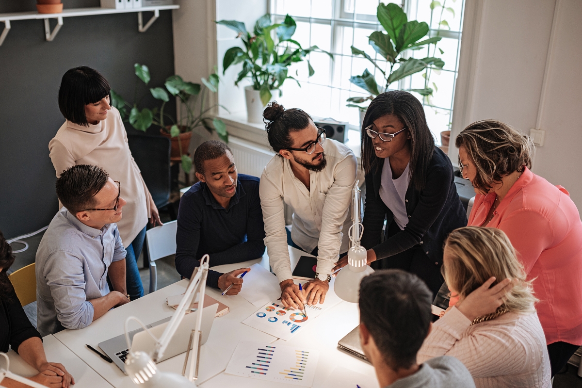 Storytelling in Marketing: Crafting Narratives that Resonate 3 A group of professionals discusses charts and data around a table in a modern office with plants and large windows, exploring storytelling in marketing to enhance their strategies.