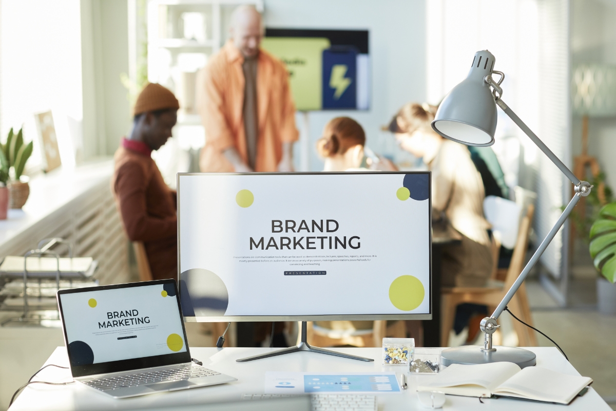 Storytelling in Marketing: Crafting Narratives that Resonate 4 Laptop and monitor display "Brand Marketing" on a desk with office supplies, while a group collaborates in the background—highlighting the power of storytelling in marketing to unite teams and engage audiences.