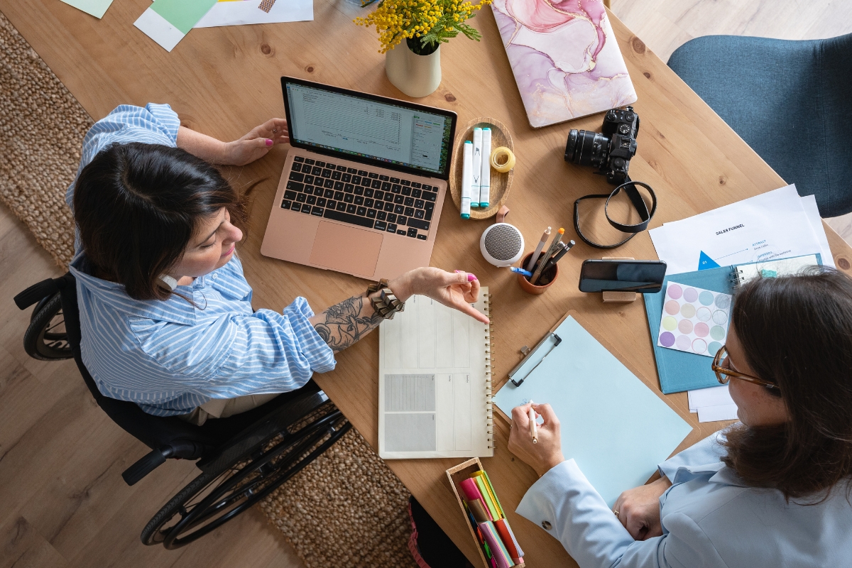 Storytelling in Marketing: Crafting Narratives that Resonate 5 Two people collaborate at a desk—with a laptop, planner, camera, documents, and art supplies—crafting creative ideas for storytelling in marketing; one person uses a wheelchair.