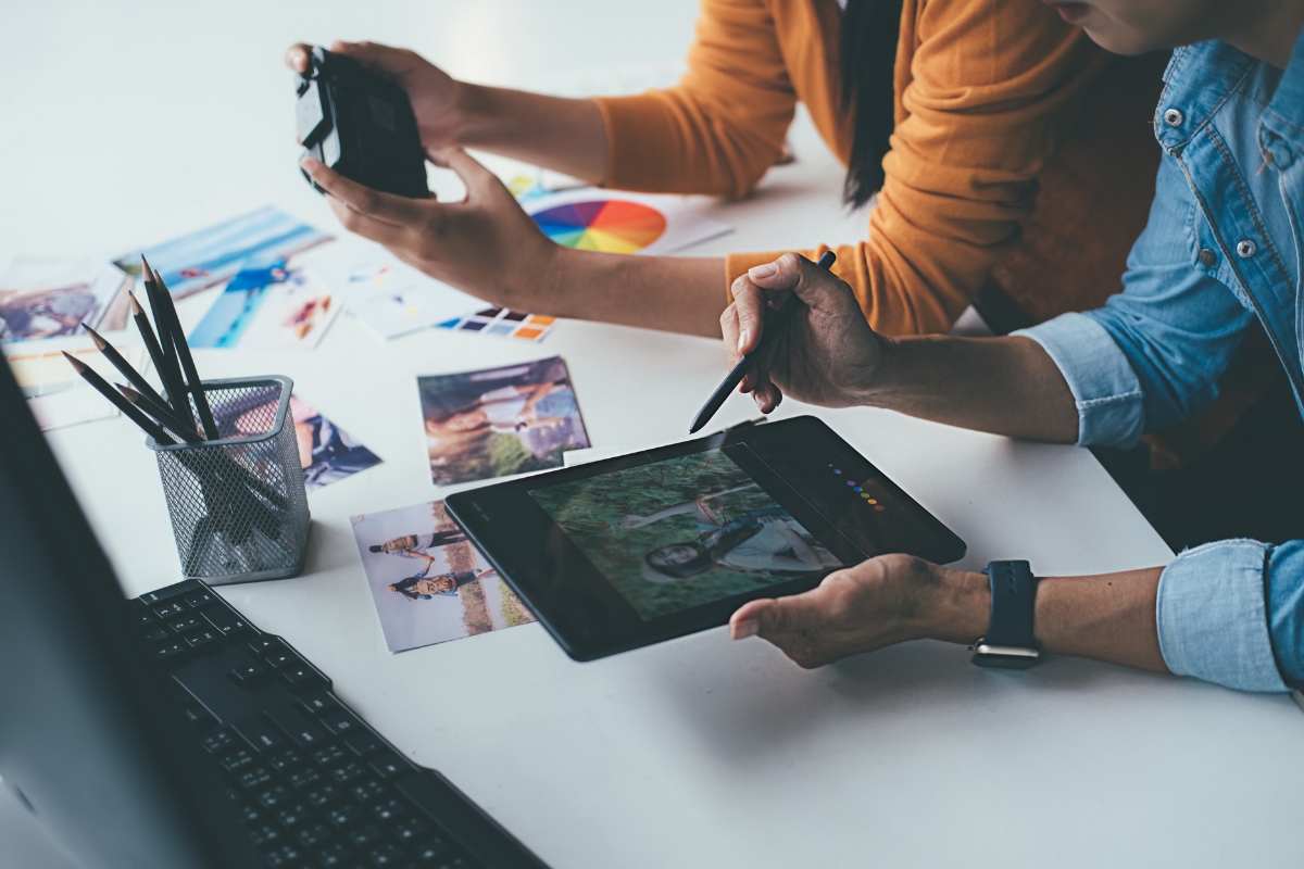 Storytelling in Marketing: Crafting Narratives that Resonate 6 Two people edit a photo on a tablet with a stylus, surrounded by printed photos, a camera, a color chart, and computer accessories—capturing the essence of storytelling in marketing for visually engaging campaigns.