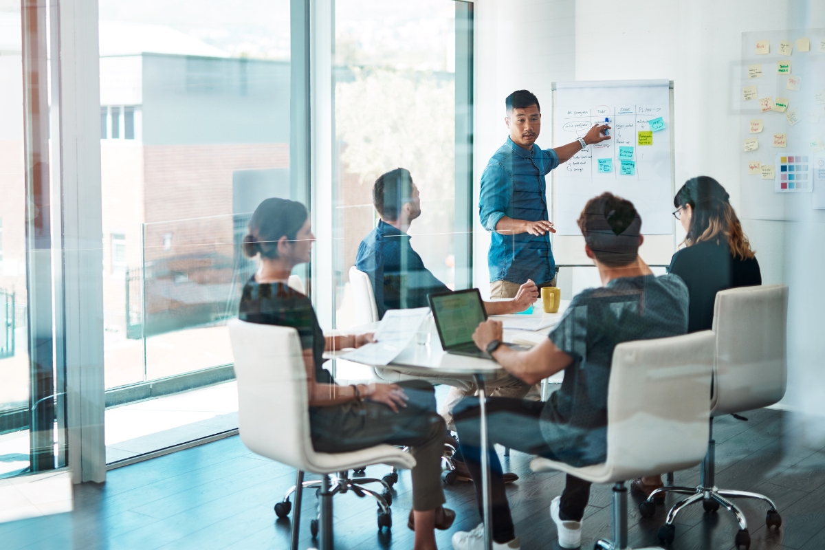 Storytelling in Marketing: Crafting Narratives that Resonate 2 Five people in a modern office meeting room, with one person standing and presenting storytelling in marketing on a whiteboard to four seated colleagues.