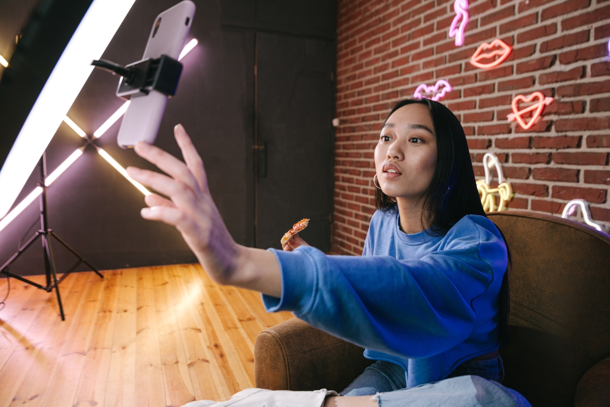 Harnessing the Power of TikTok for Business Marketing 2 A person in a blue sweater holds pizza and reaches toward a phone on a tripod, possibly filming Tiktok for business marketing, seated in a room with neon wall lights and exposed brick.