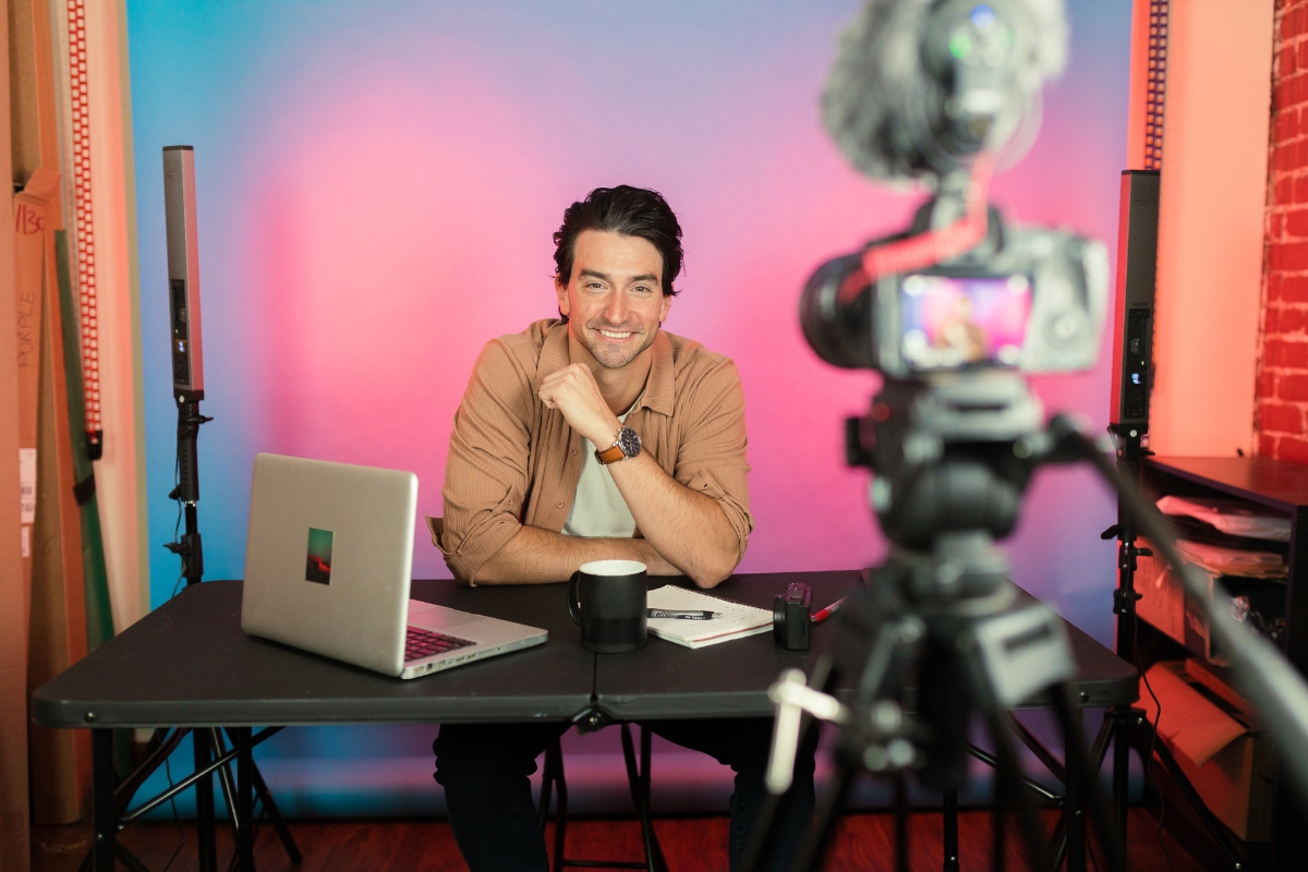 Harnessing the Power of TikTok for Business Marketing 6 A man sits at a desk with a laptop, mug, notepad, and camera, recording a Tiktok for business marketing video in a studio with a colorful background.